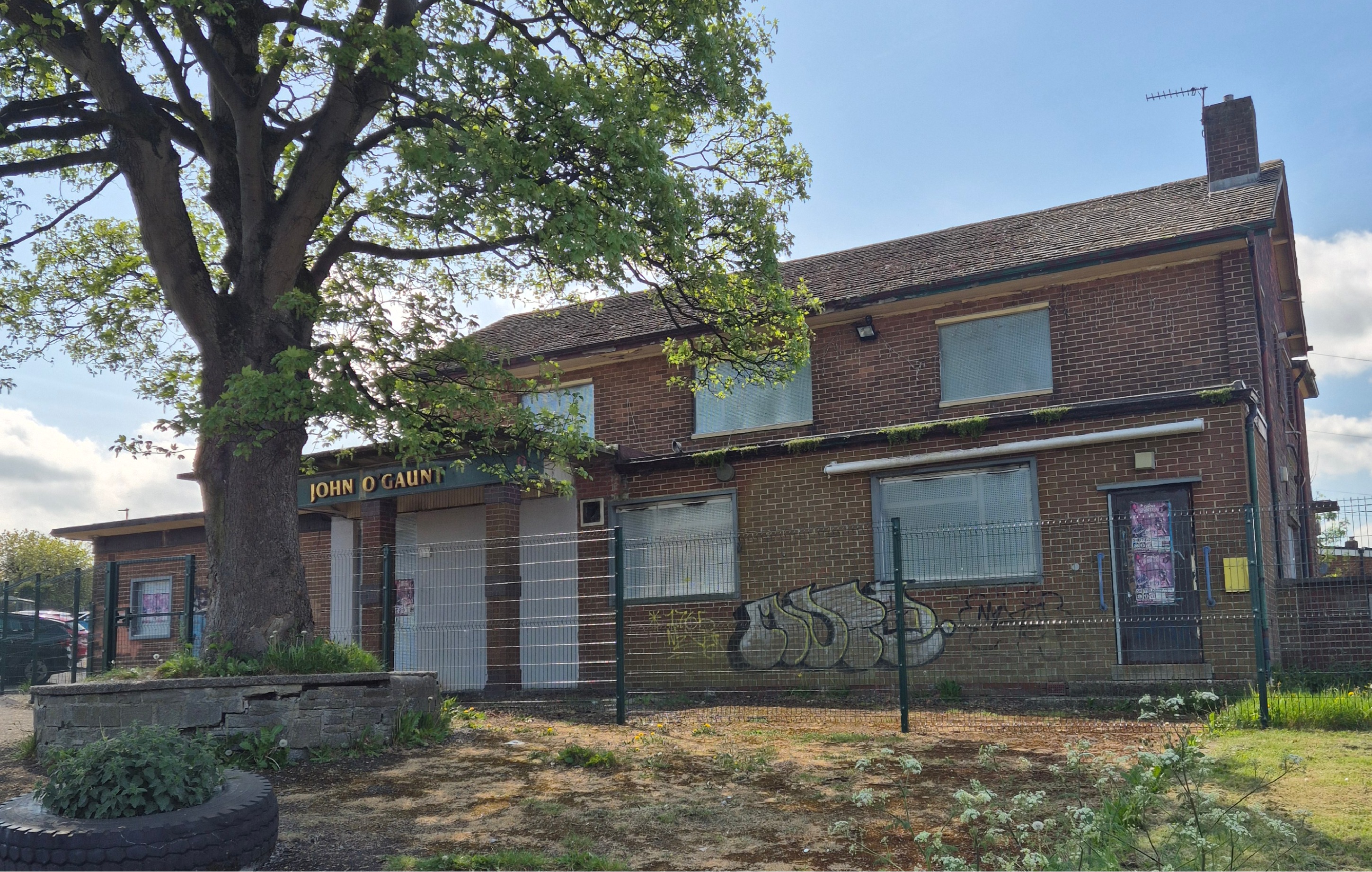 The exterior of the John O'Gaunt pub with boarded windows and fencing around it. Tree and grass in foreground with blue clear skies.