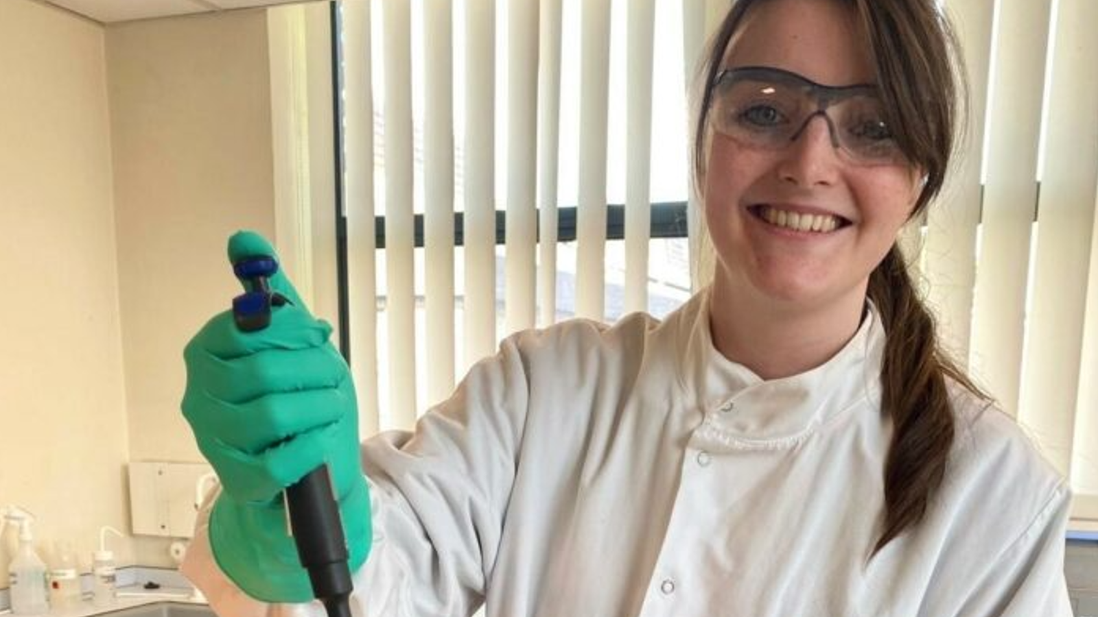 A person in a white lab coat and green gloves holds a pipette, set against a background of vertical blinds and a laboratory setting.