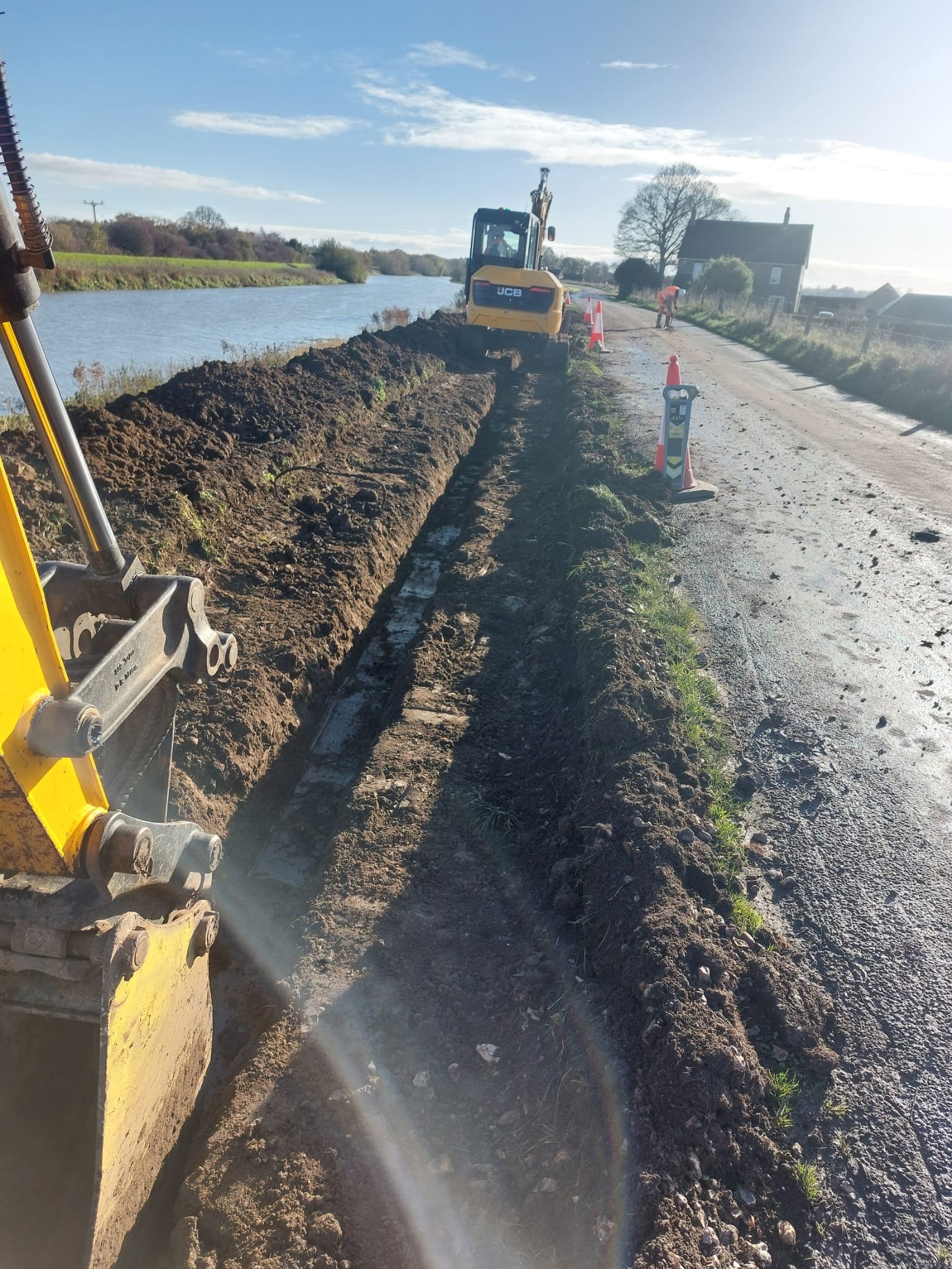 Excavation works taking place along a river embankment, with a digger digging a trench and traffic cones marking the work area on the road.