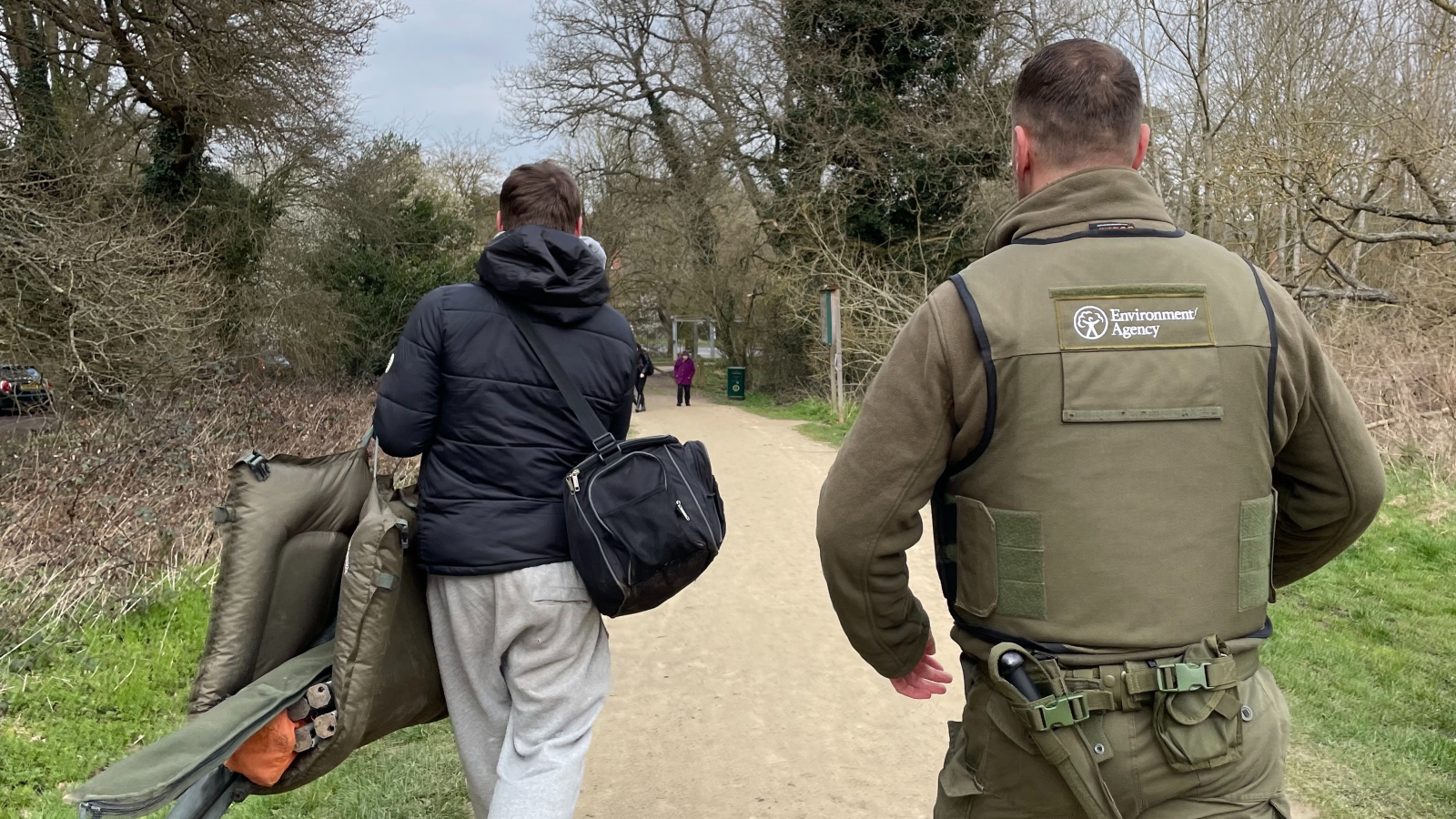 Two men walk along a path in a wooded area; one carries a chair, while the other wears an Environment Agency uniform.