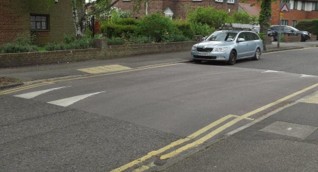 Image shows a bus-friendly road hump that stretches all the way across the road from kerb to kerb. There are double yellow lines at either side of the road and tactile paving on the pavement to indicate the crossing. 
