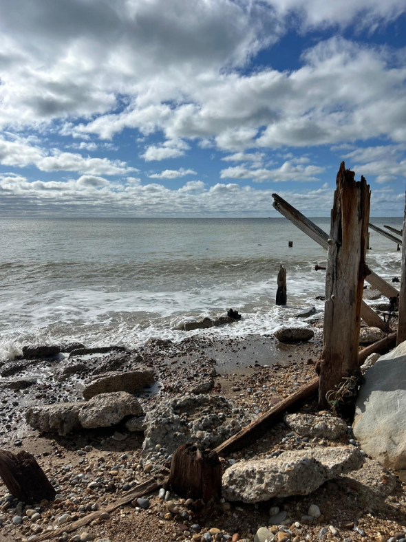 Photo shows a the coast with blue sky and clouds with the shoreline in the foreground with rocks and a wooded post