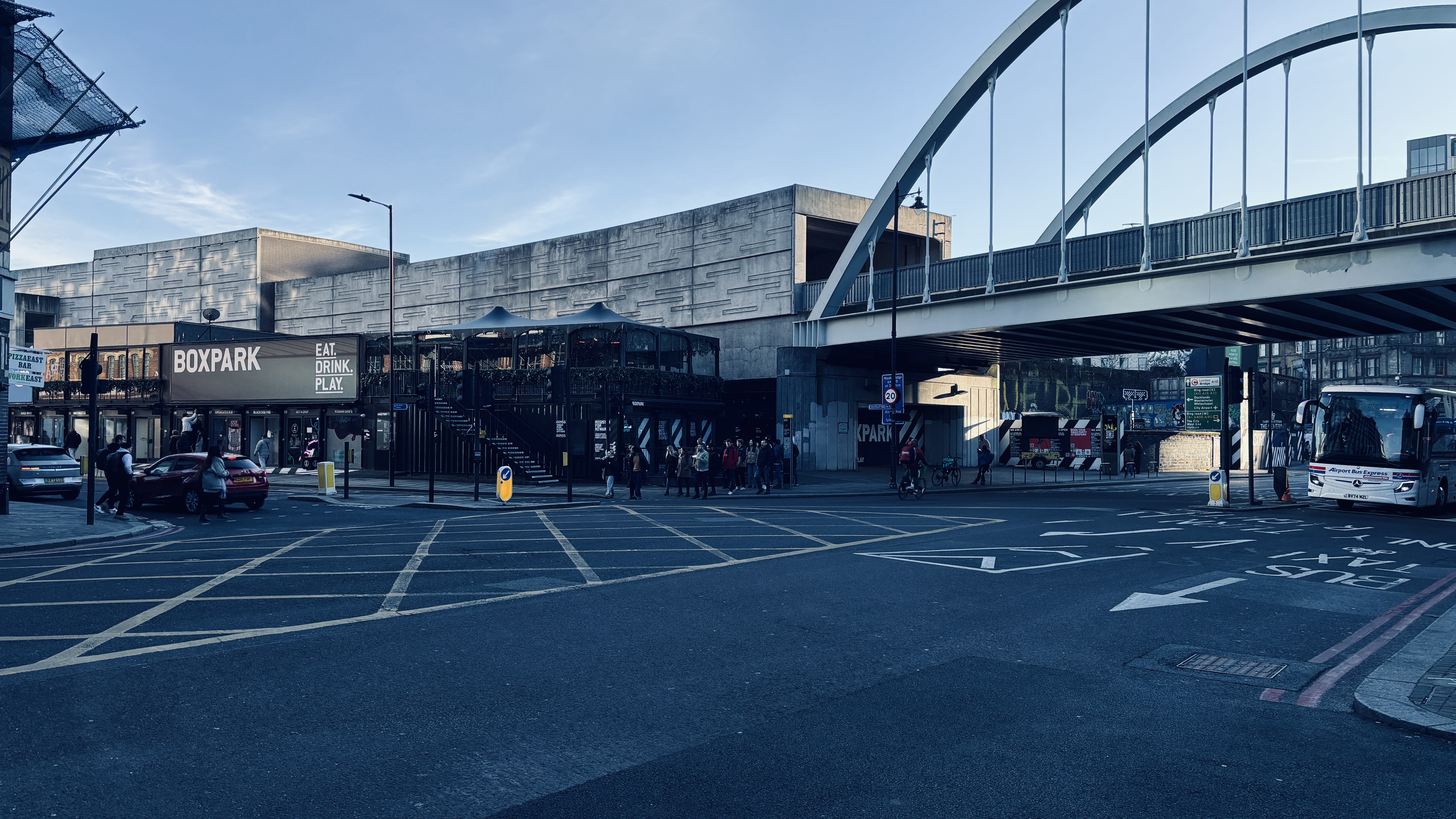 Urban street with yellow grid markings, a pedestrian crossing, arched bridge, some pedestrians and parked cars