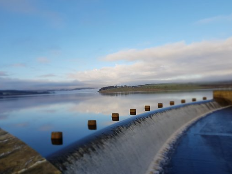 Calm water reflecting the sky alongside a stone barrier with wooden posts, under a blue sky and wispy clouds.