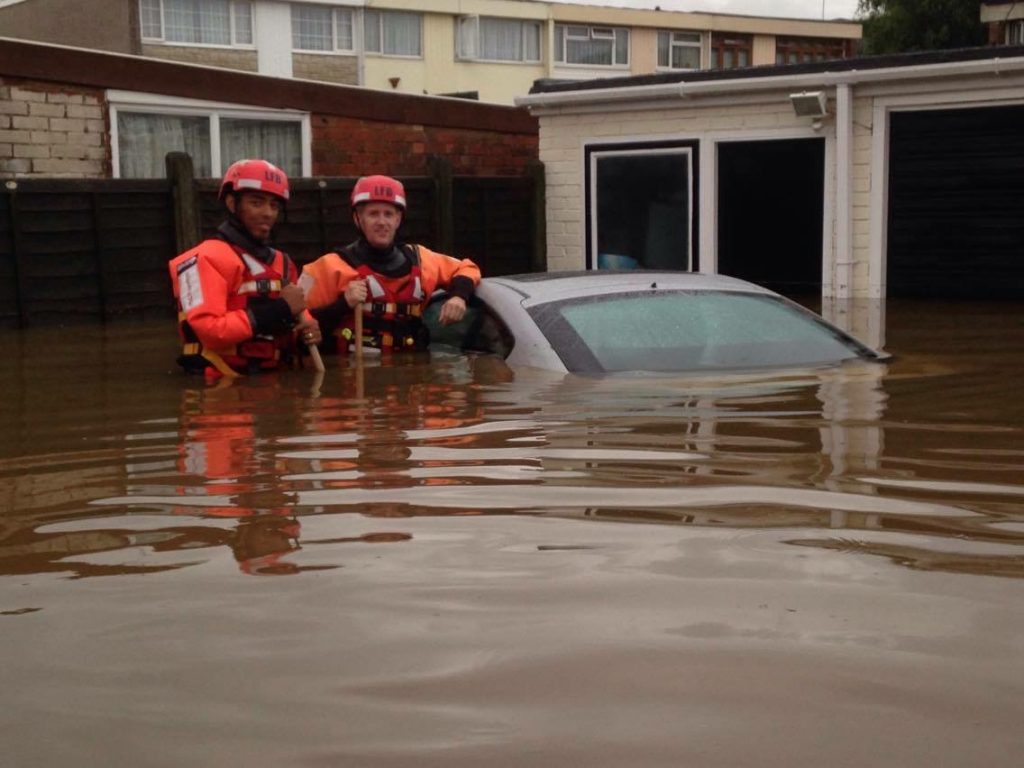 A picture showing two emergency responders standing in flood water. The flood water is above their waist and has almost submerged the car they are leaning against.