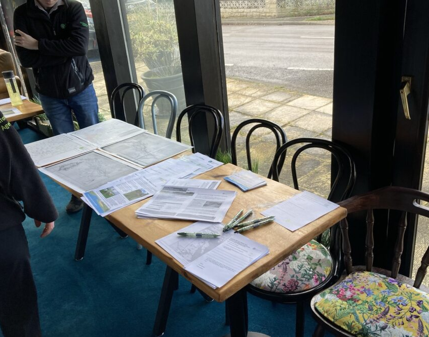 A table displayed with project information including various maps, newsletters, briefing notes, and feedback forms. Two members of Environment Agency staff can be seen manning the table. 