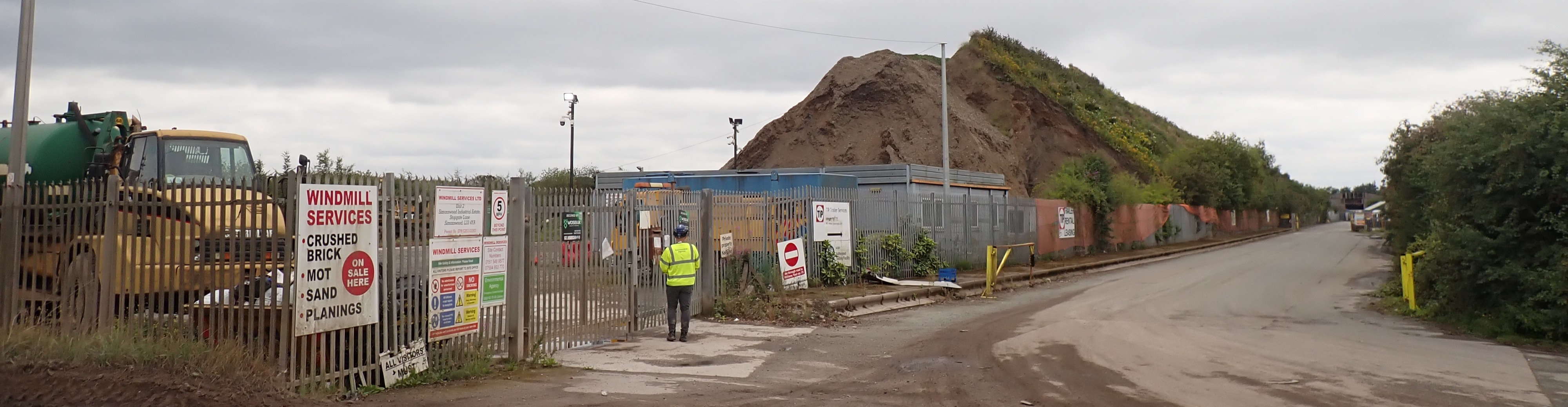 EA Officer outside gates of Windmill site, site vehicles and waste pile visible 