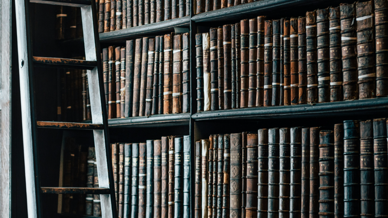 A close-up of a wooden bookshelf filled with vintage leather-bound books, alongside a rustic ladder for reaching higher shelves.