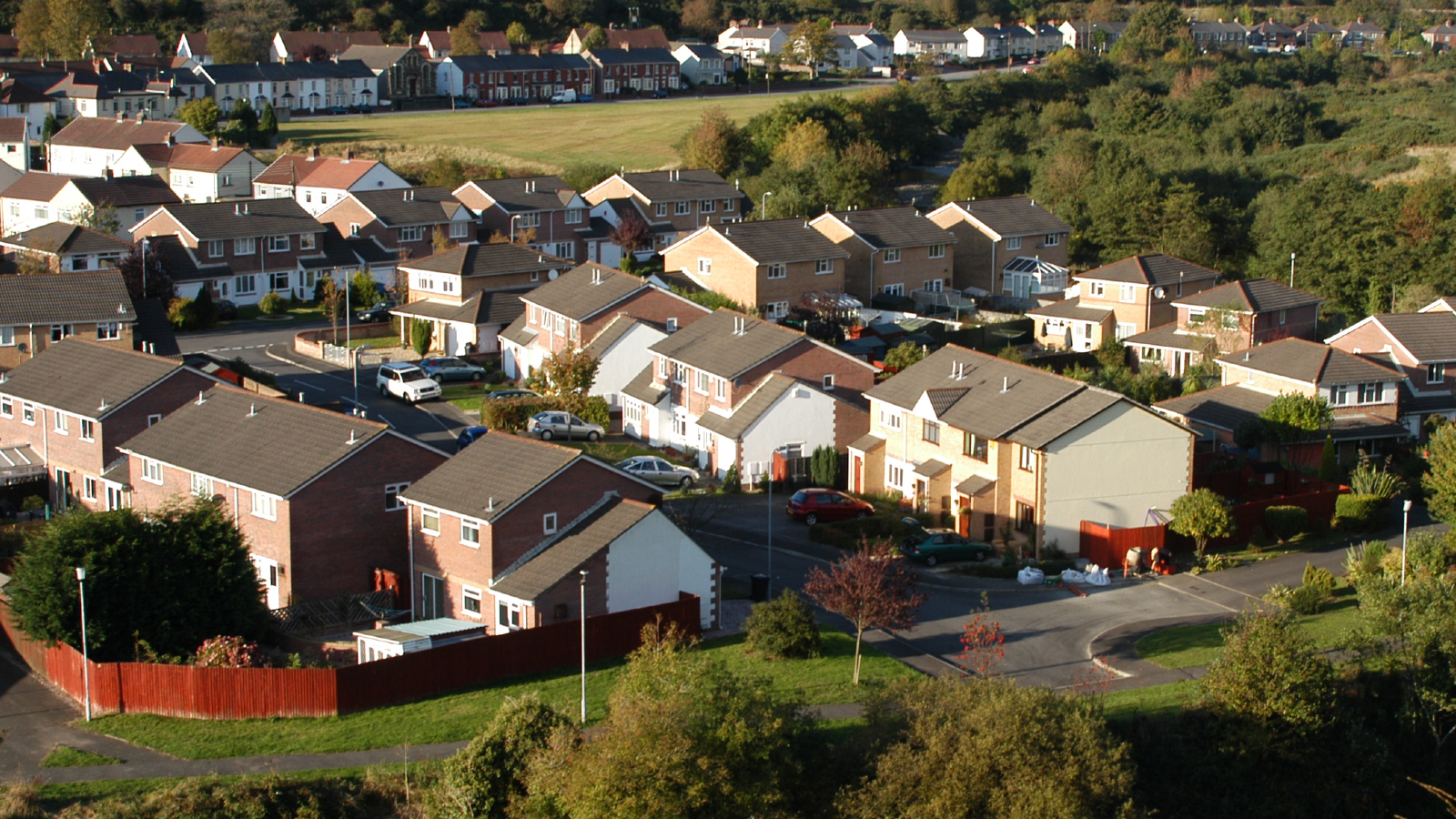 Aerial view of a suburban neighborhood with houses, green lawns, and parked cars, surrounded by trees and open fields.