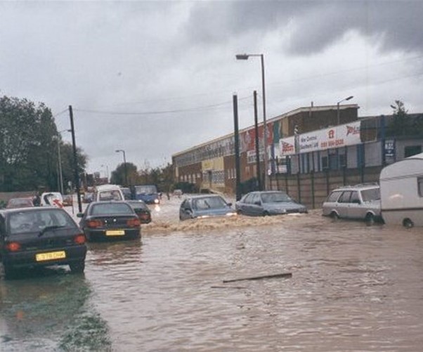 Picture showing a flooded road with parked cars. One car is driving through the flood water
