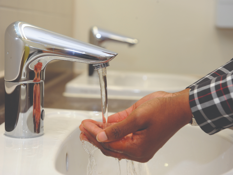 A person with a checkered shirt washes their hands under a modern chrome faucet, with water flowing into a sink.