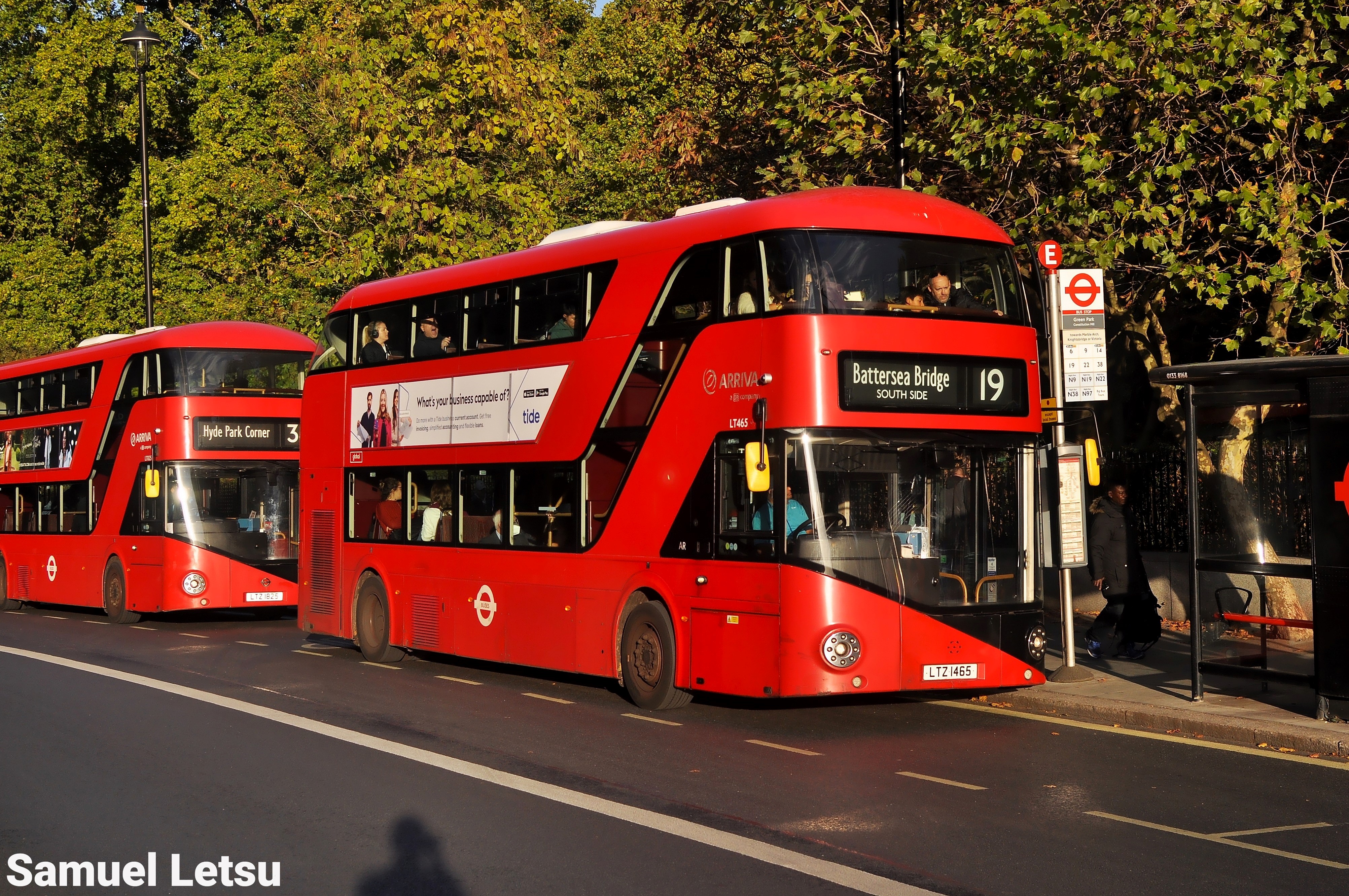 Two red double-decker London buses at a bus stop, with the front bus on route 19 to Battersea Bridge and the second bus on route 3 to Hyde Park Corner, against a backdrop of trees and a bus shelter