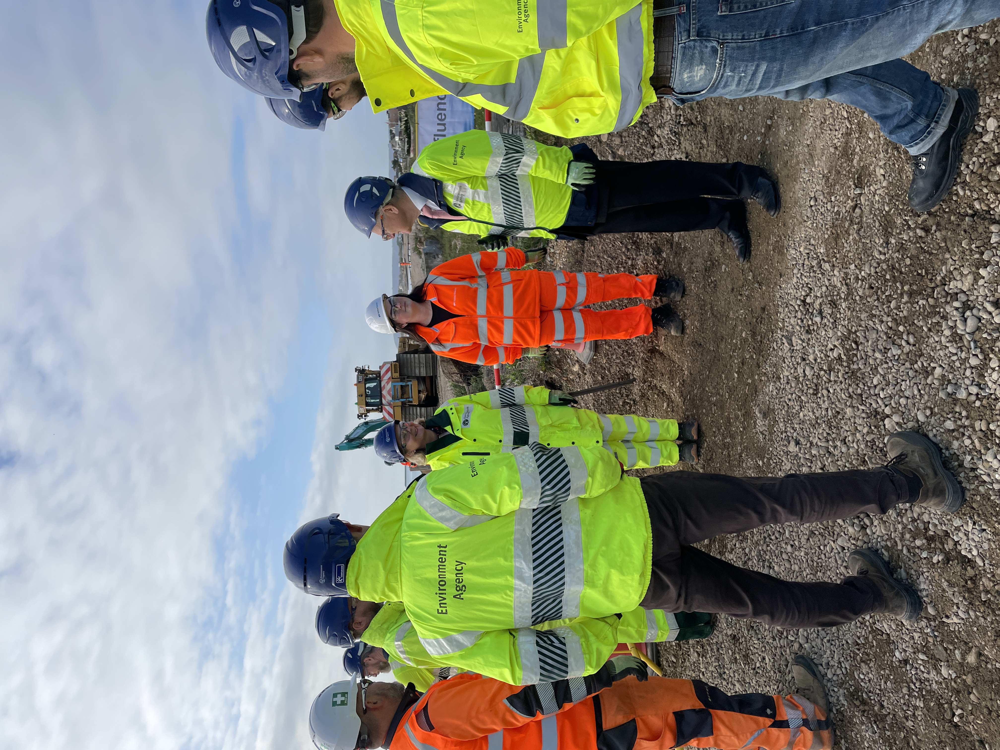 People in high-visibility jackets and hard hats standing on gravel at Pevensey Bay beach with construction equipment in background.