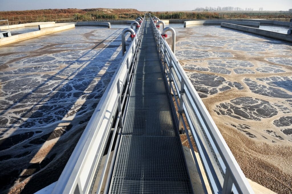 A walkway over a water treatment facility with foamy wastewater and industrial pipes, surrounded by greenery under a clear sky.
