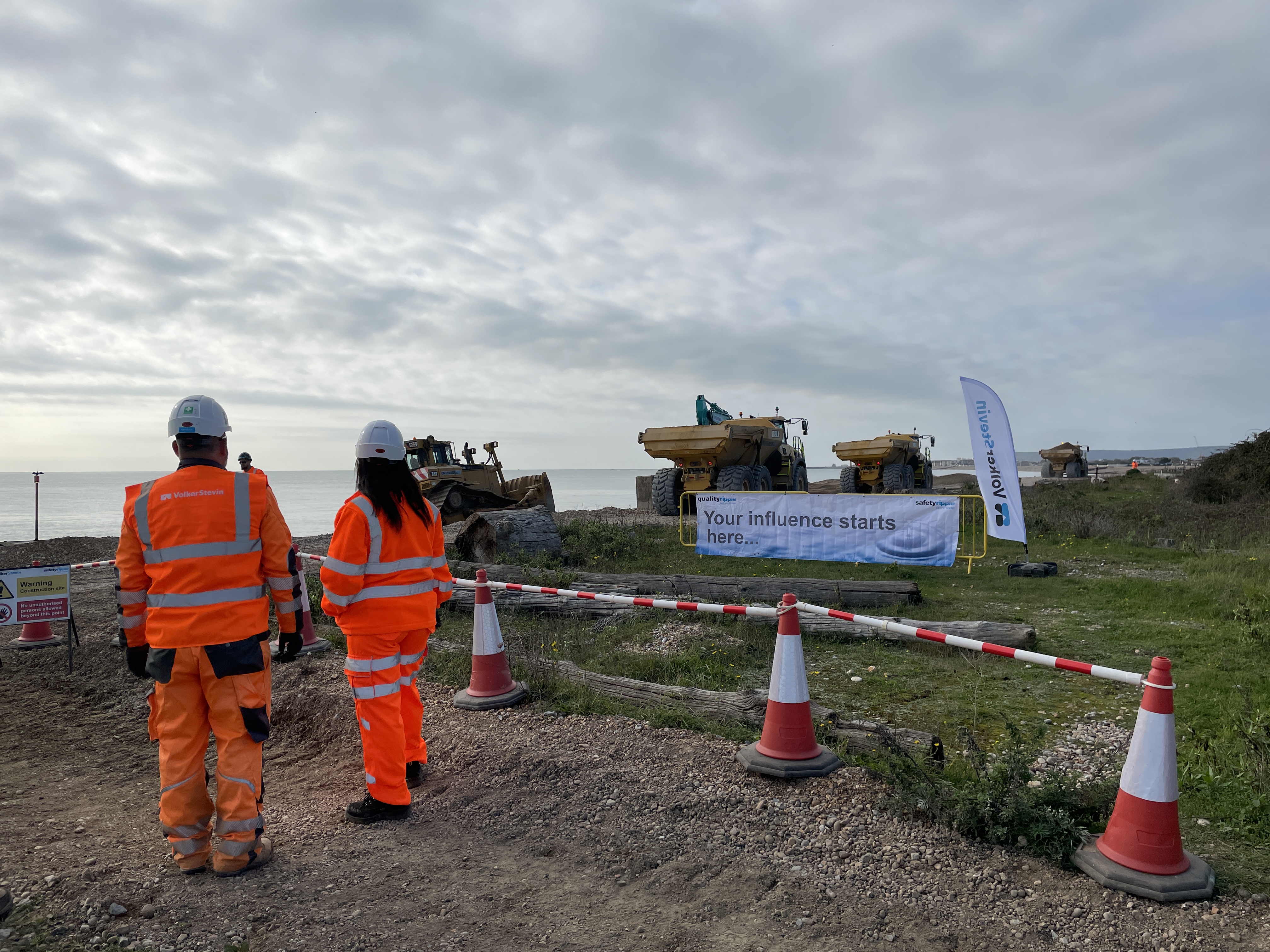 People in high-visibility jackets and hard hats observe coastal construction at Pevensey beach with vehicles behind.
