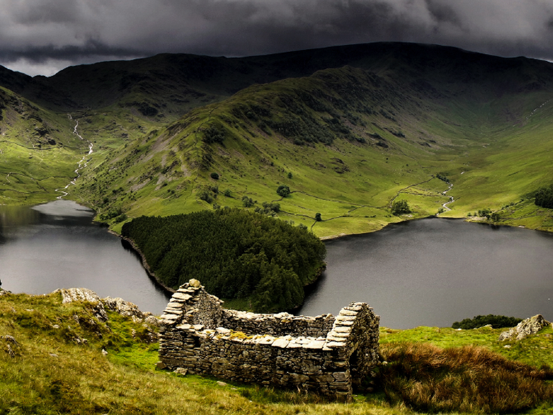 A stone ruin overlooks lush green valleys, reflecting lakes, and dramatic mountains under a brooding, cloud-filled sky.