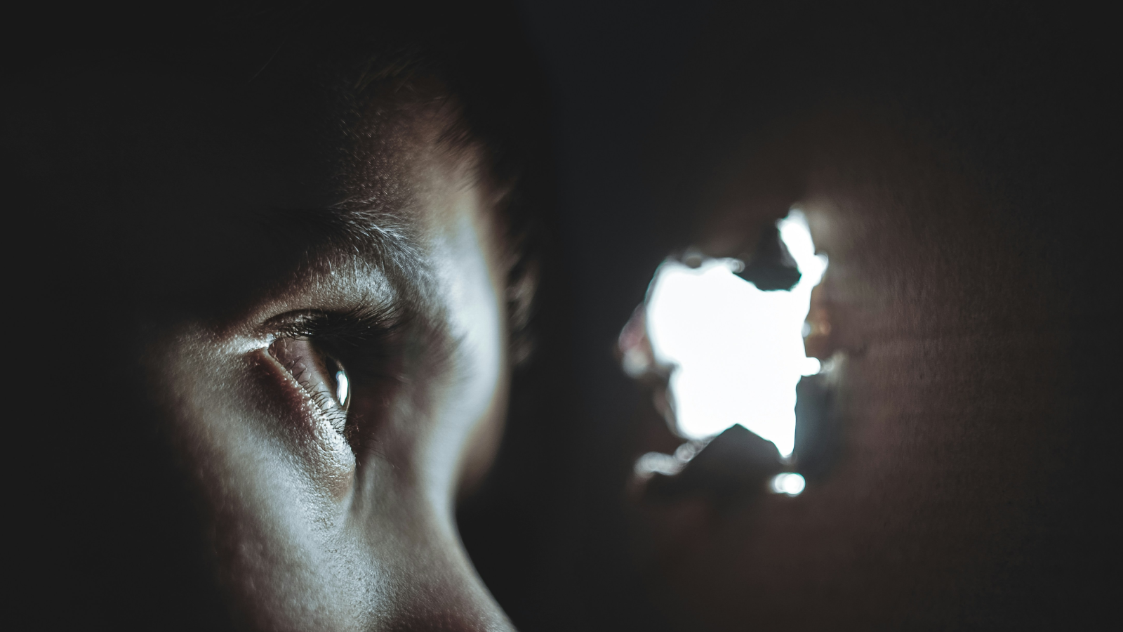 A close-up of an eye peering through a hole in a dark wall, with light streaming in.