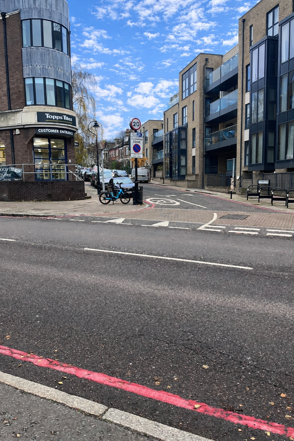 Bishop’s Road junction with modern apartments and a shop on the corner, parked cars, a bicycle, and a visible 20 mph speed‑limit sign under a bright blue sky