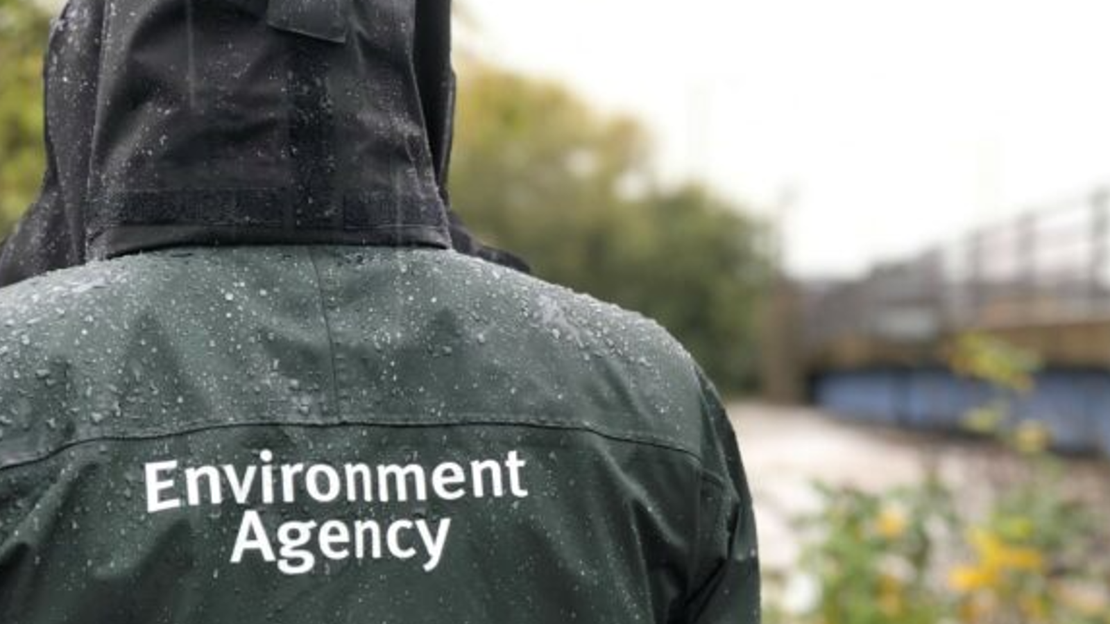 A person wearing a rain jacket with "Environment Agency" printed on the back, standing outdoors in a wet, rainy environment.