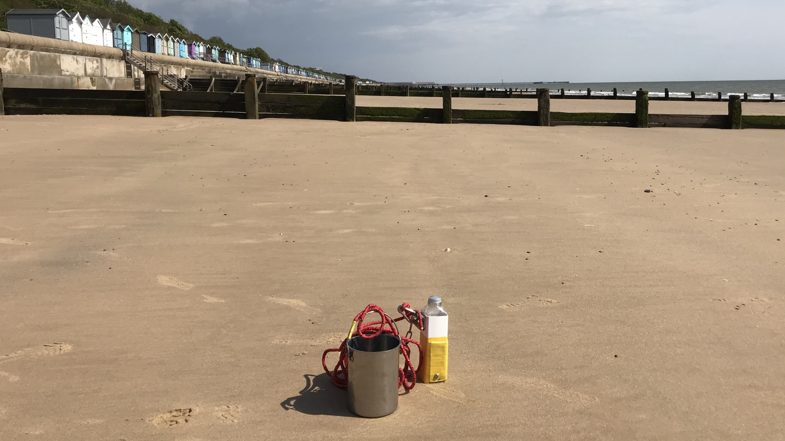 A metal bucket, red rope, and a bottle sit on a sandy beach with colorful beach huts in the background and the ocean in the distance.