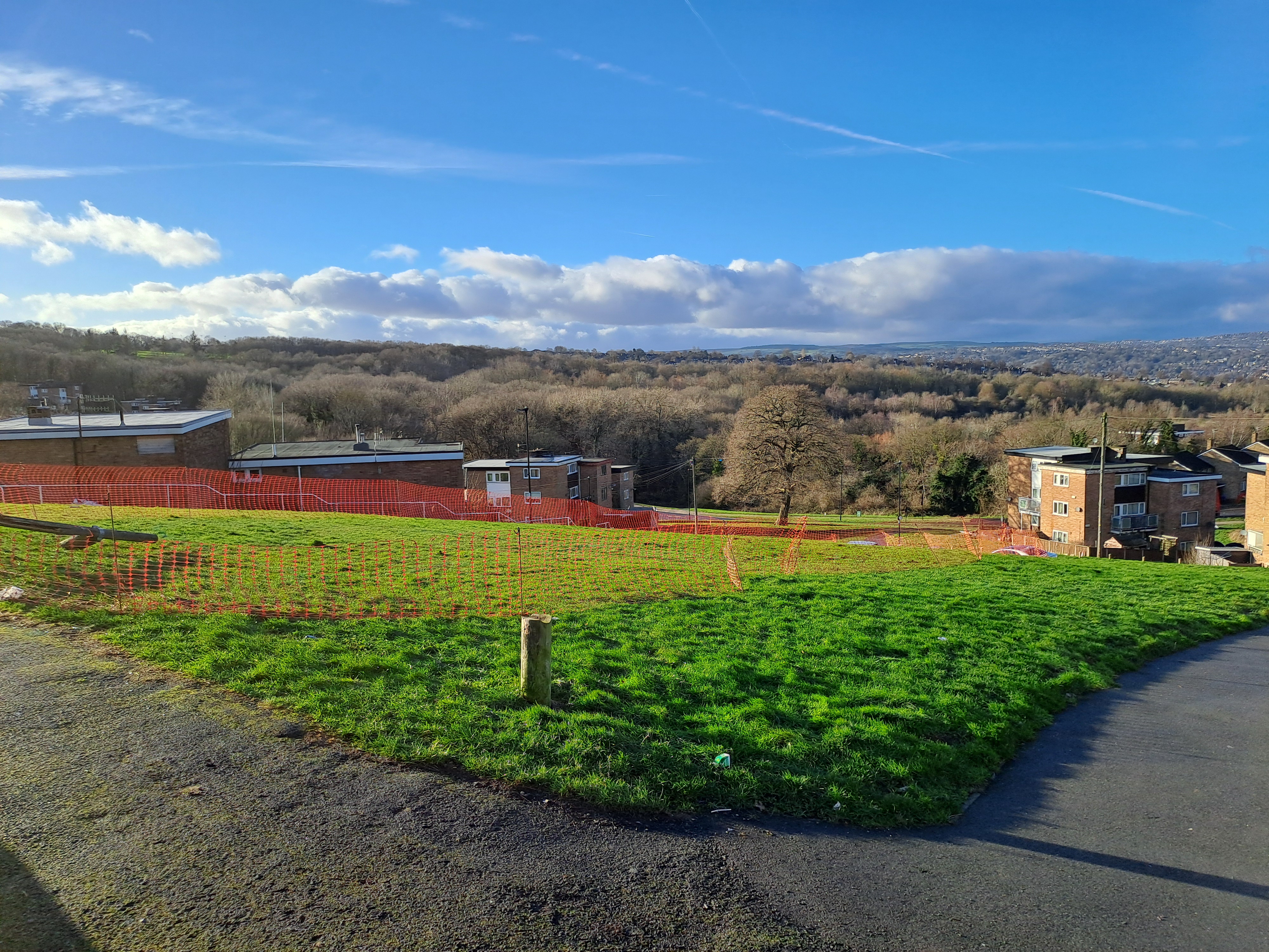 A view of the large grassed area at Spotswood on a sunny day. Orange safety net fencing has been installed around the perimeter. There are trees in the distance and blue skies above.