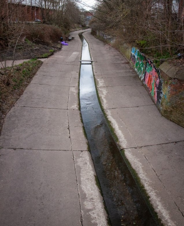 A view of a 50 metre length of concreted channel with low sides. The channel is extremely clean, bordering on spotless down this entire stretch, and the water flowing in the central summer channel looks clear.