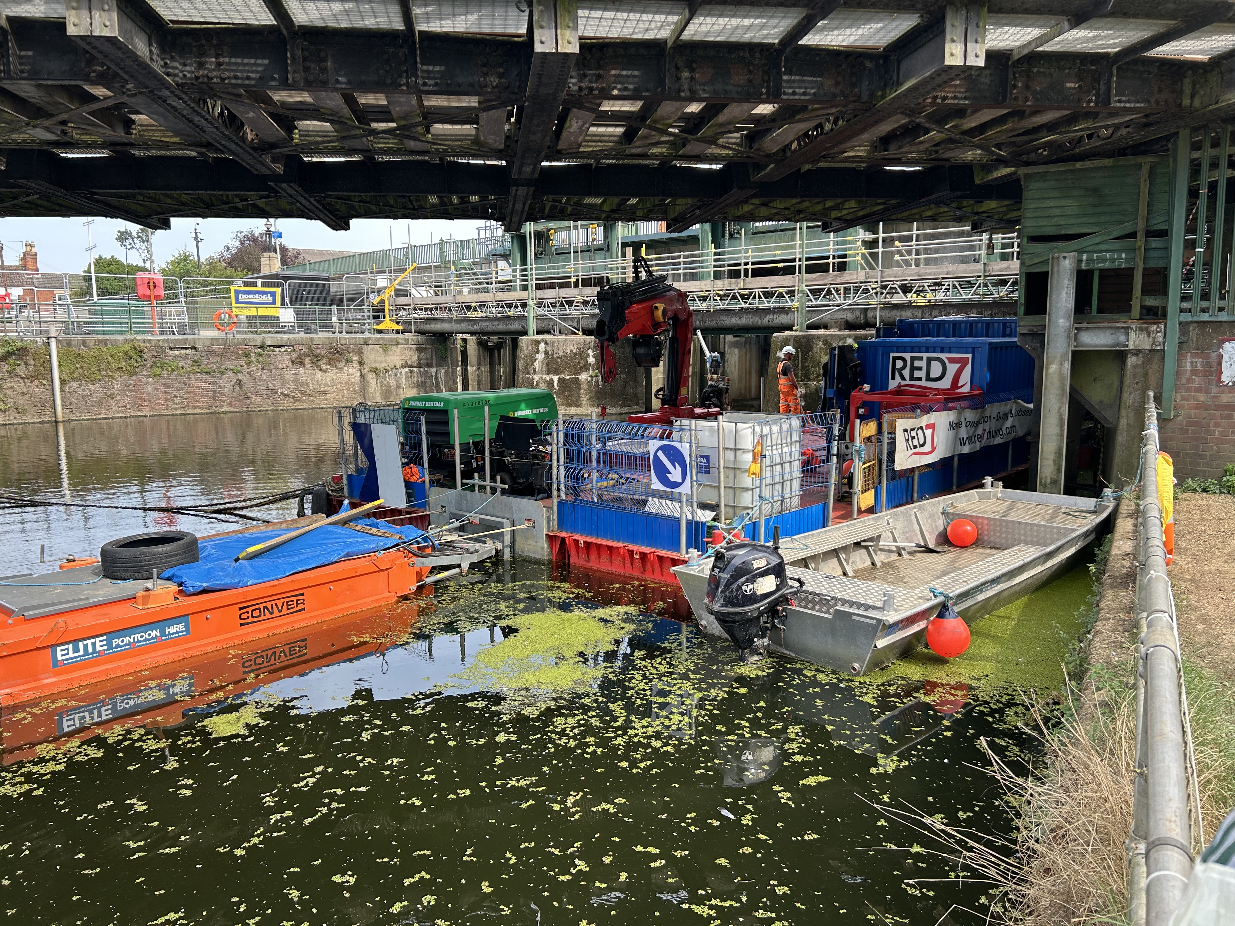 Multiple work boats and floating platforms are positioned beneath a metal bridge in the river channel. Workrs in protective clothing stand on the barge with a crane. A small metal boat is tied up alongside, green plant material can be seen floating on the surface of the water. Fencing, scaffolding and signs are visible along the edge of the river and bridge structure. 