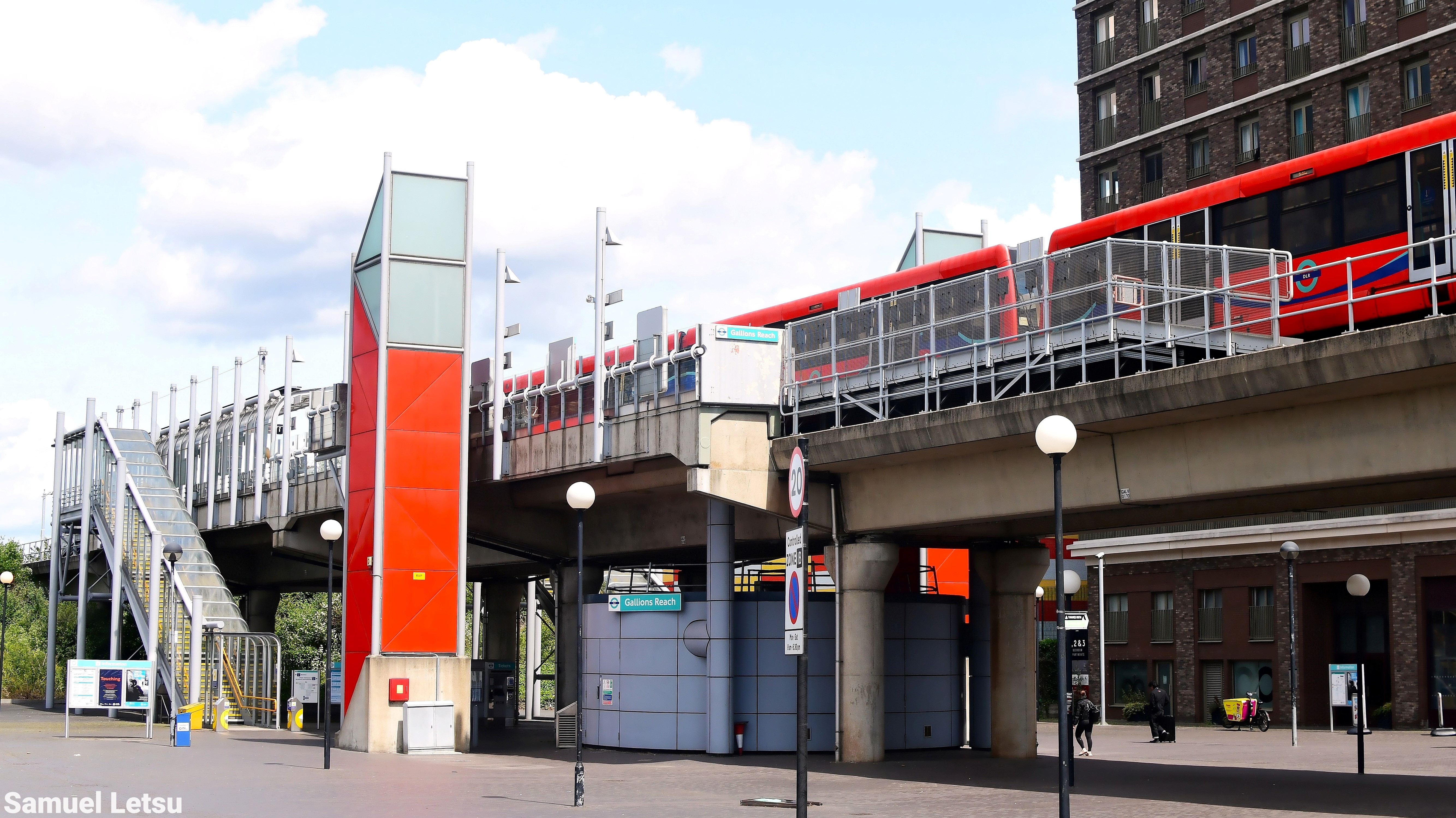 DLR train at Gallions Reach station on an elevated platform with surrounding buildings and pedestrian access stairs