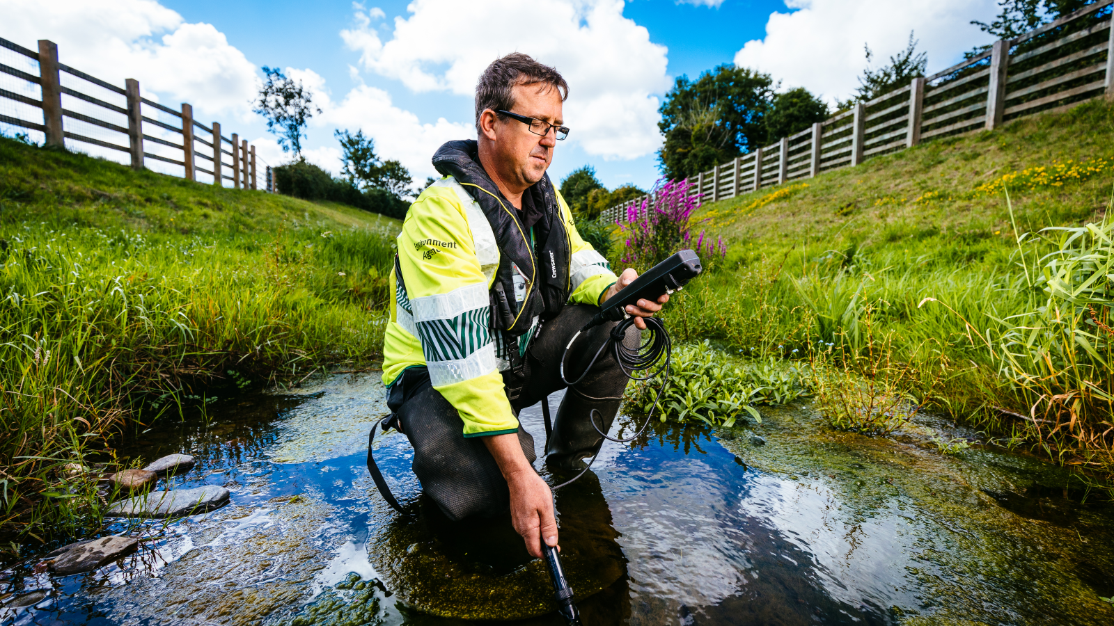A person in a fluorescent jacket kneels in a stream, holding a water testing device, surrounded by lush greenery and flowers.