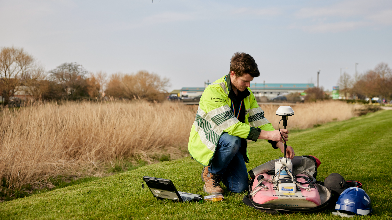 A technician in a reflective jacket kneels on the grass, working with equipment next to a laptop and surveying gear by a waterway.