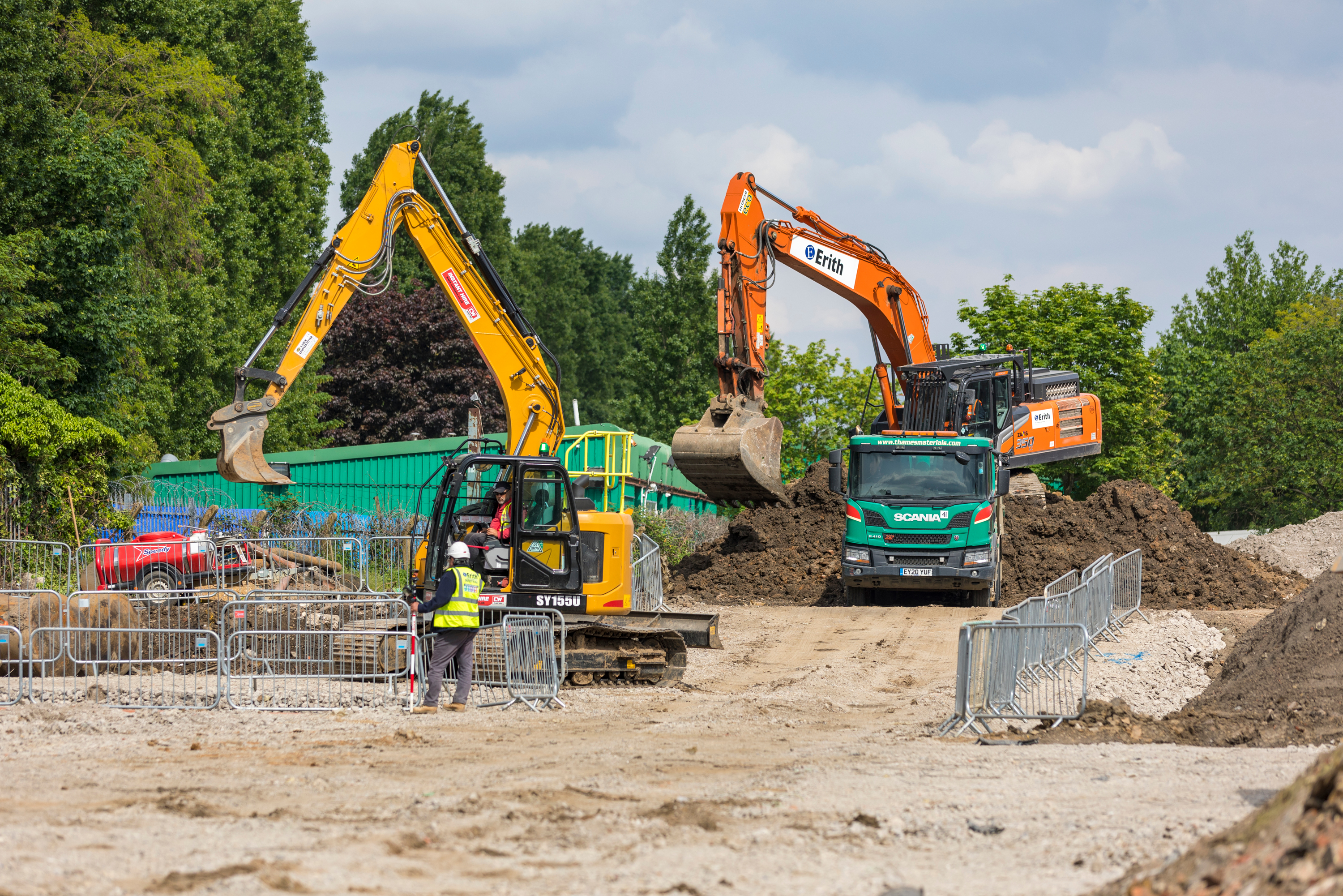 Construction site with two excavators digging and loading soil into a lorry, with workers and safety barriers visible