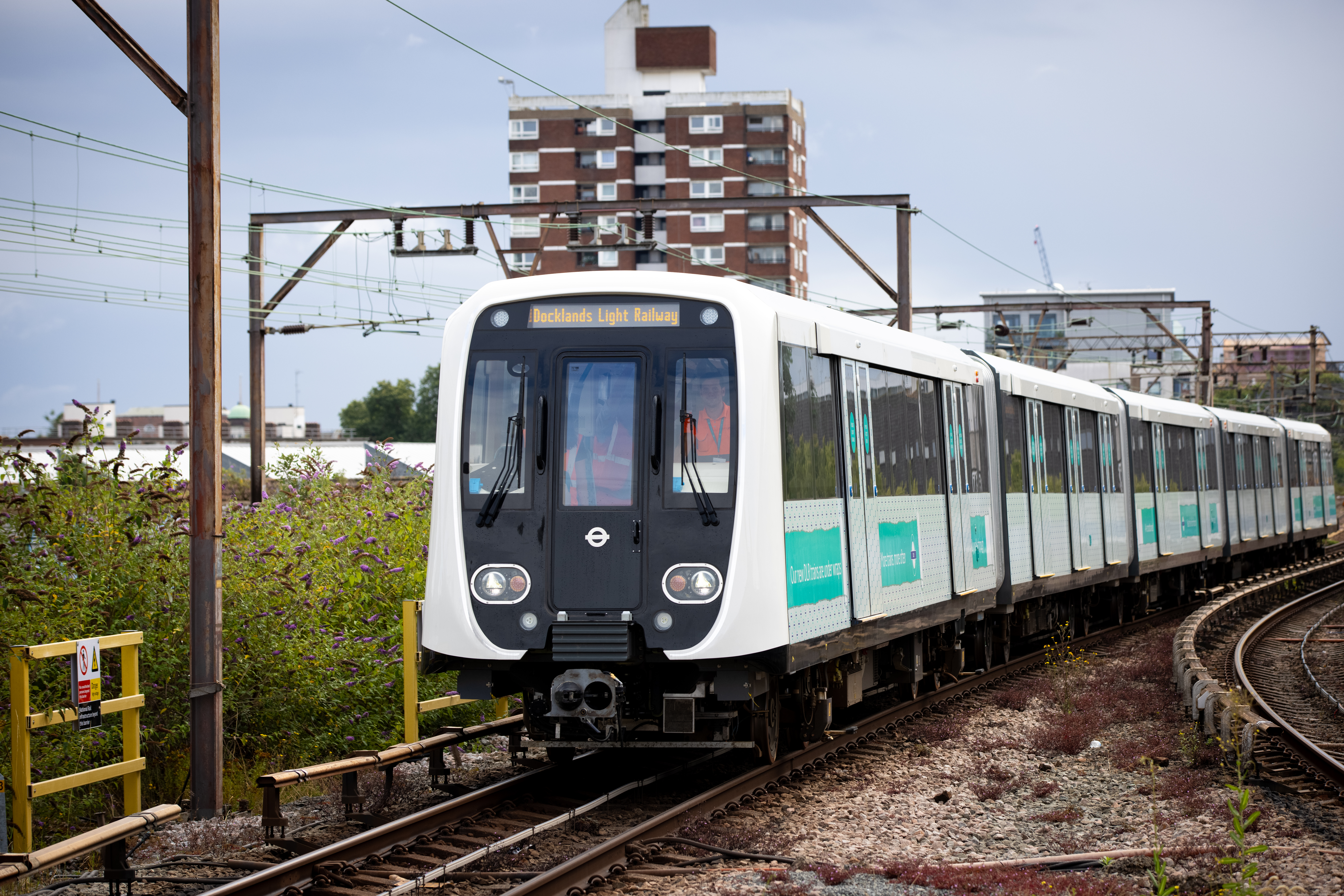 New DLR train travelling on curved tracks through an urban area with overhead lines and residential buildings in the background