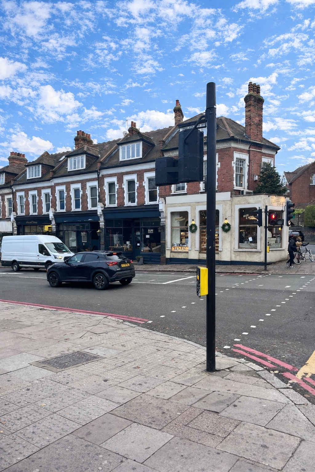 Archway Road junction showing shops and parked vehicles, including a corner bakery and pedestrians on the pavement, under a bright blue sky with scattered clouds