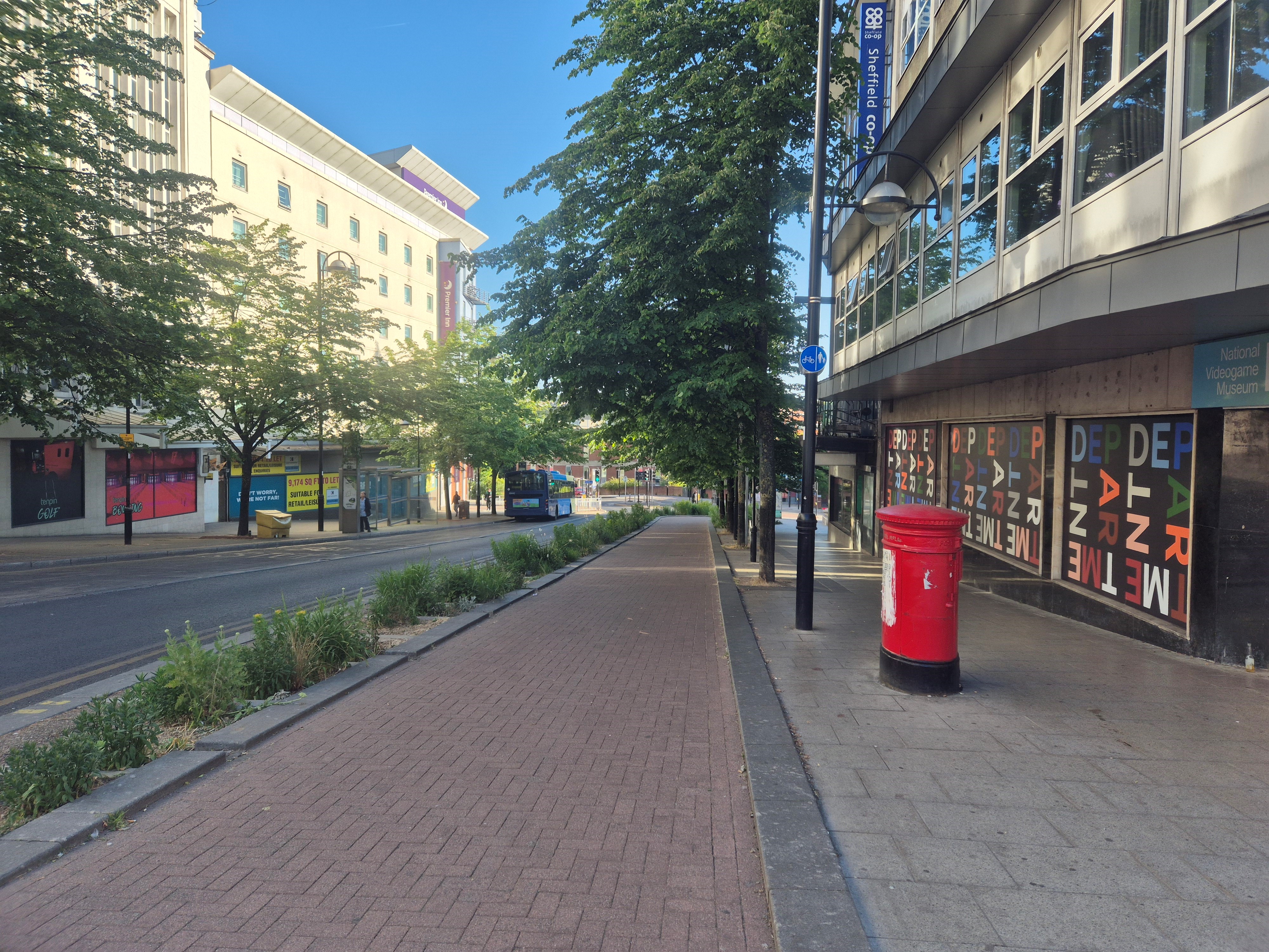 A city centre street with a red brick protected cycle route. there are trees and buildings on either side of the road, planting between the carriageway and the cycle route and a red pillar box stands to the right. A bus can be seen in the distance and blue sky is overhead.