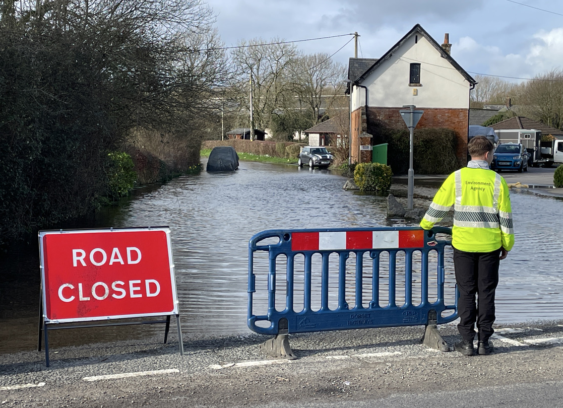 Environment Agency Officer standing by barrier and 'road closed' sign blocking flooded road