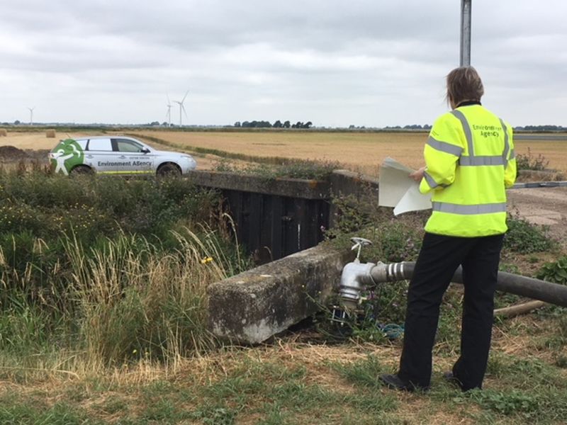 A woman in a high-visibility jacket inspects a water outlet near a field, with an Environment Agency vehicle and wind turbines in the background.