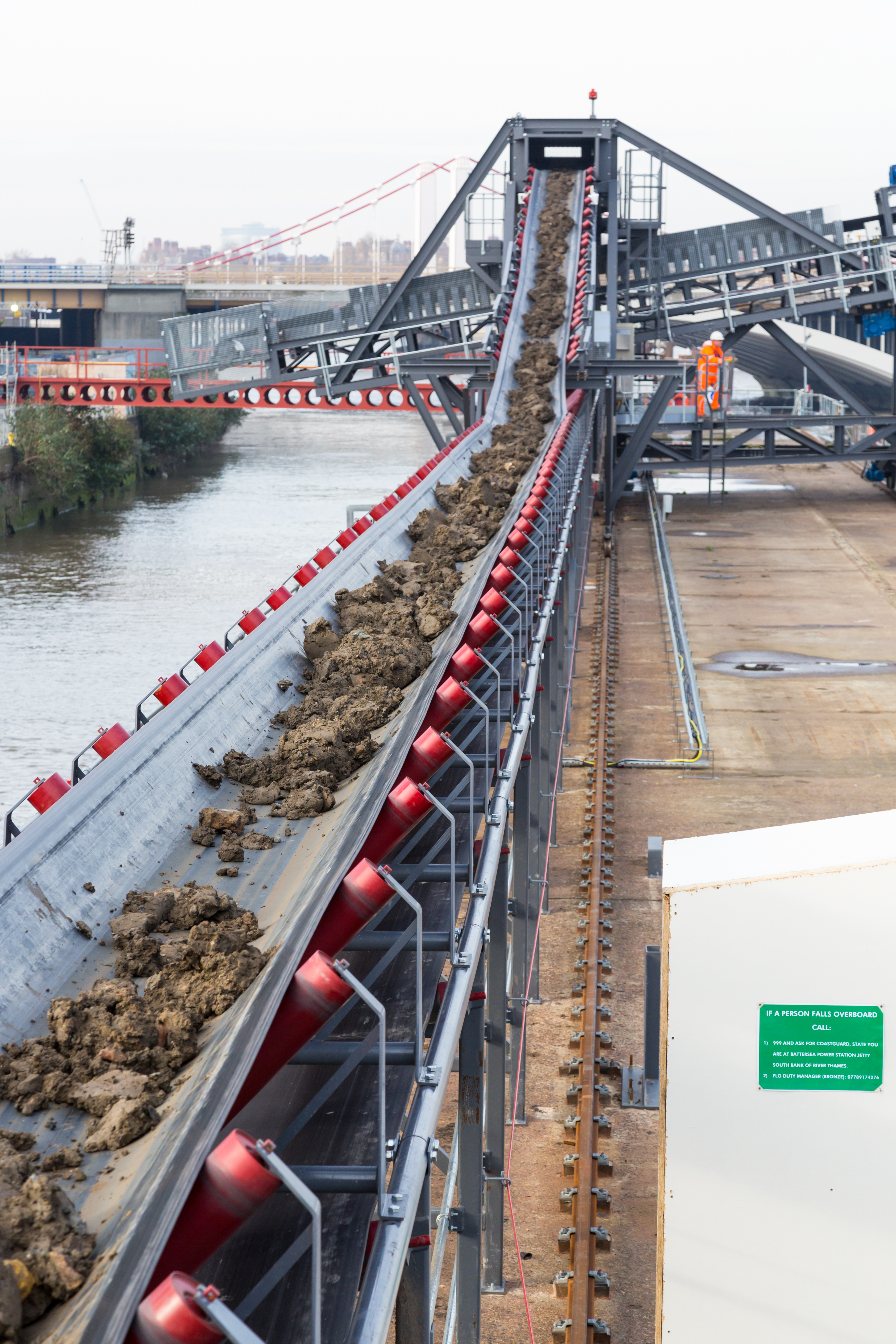Conveyor belt transporting excavated soil at a riverside construction site with industrial equipment and workers in the background