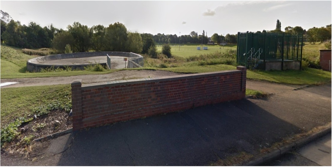 A photo of Willow Brook Park. The park is situated in a large hollow or bowl, several metres deep and about 200 metres across in both directions. A football pitch is visible within the park and it's a bright sunny day. A large circular concrete spillway that looks like it wouldn't be out of place in a skate park, is situated in the near corner of the park a few metres above the ground level of the park.