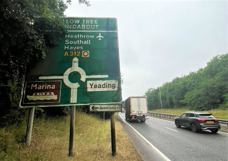 A photo of the approach to Willow Tree Roundabout, showing a large road sign and vehicles driving on the road.