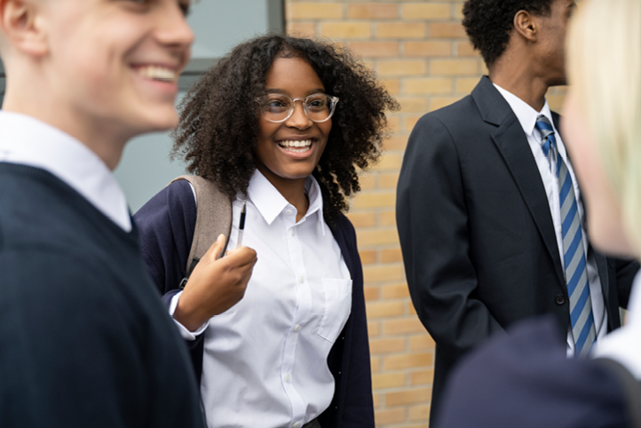Four students are dressed in school uniform standing next to a brick wall. In the foreground, a girl is laughing surrounded by three others.