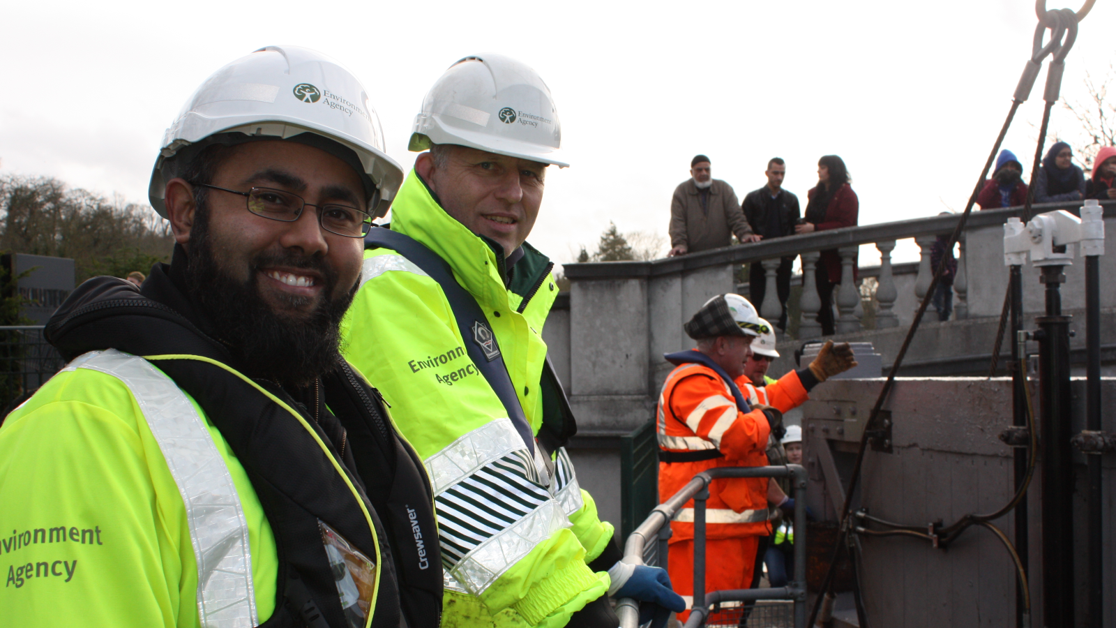 Two workers in reflective yellow jackets and hard hats survey a scene, while a third in an orange jacket operates machinery nearby.