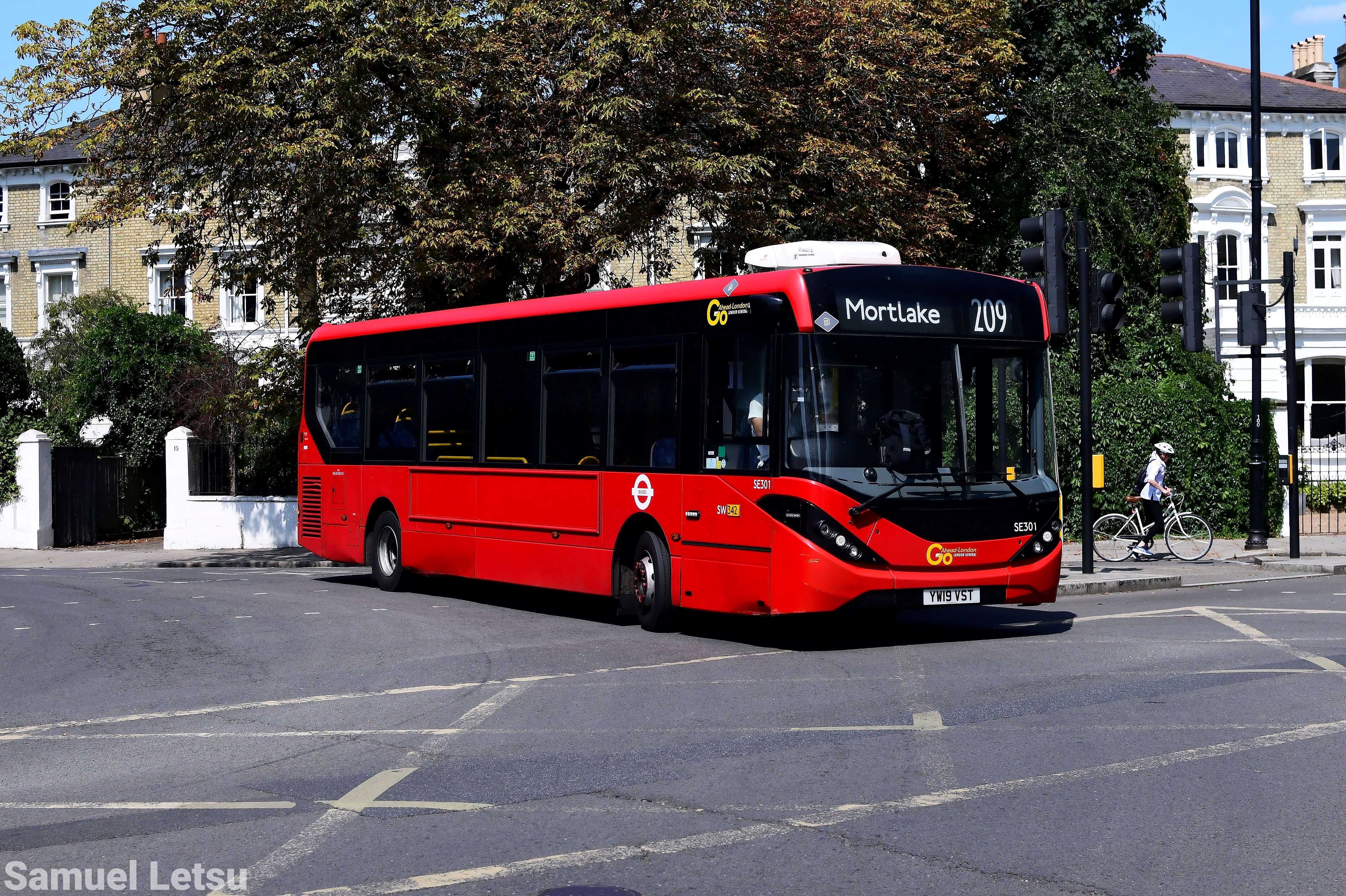 Single-decker red bus on route 209 to Mortlake at an intersection, with a cyclist in the background and residential buildings surrounded by trees