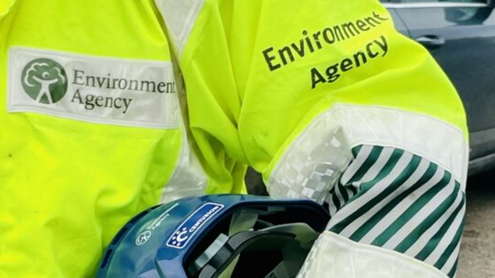 A close-up of a person in a high-visibility jacket labeled "Environment Agency," holding a blue helmet.