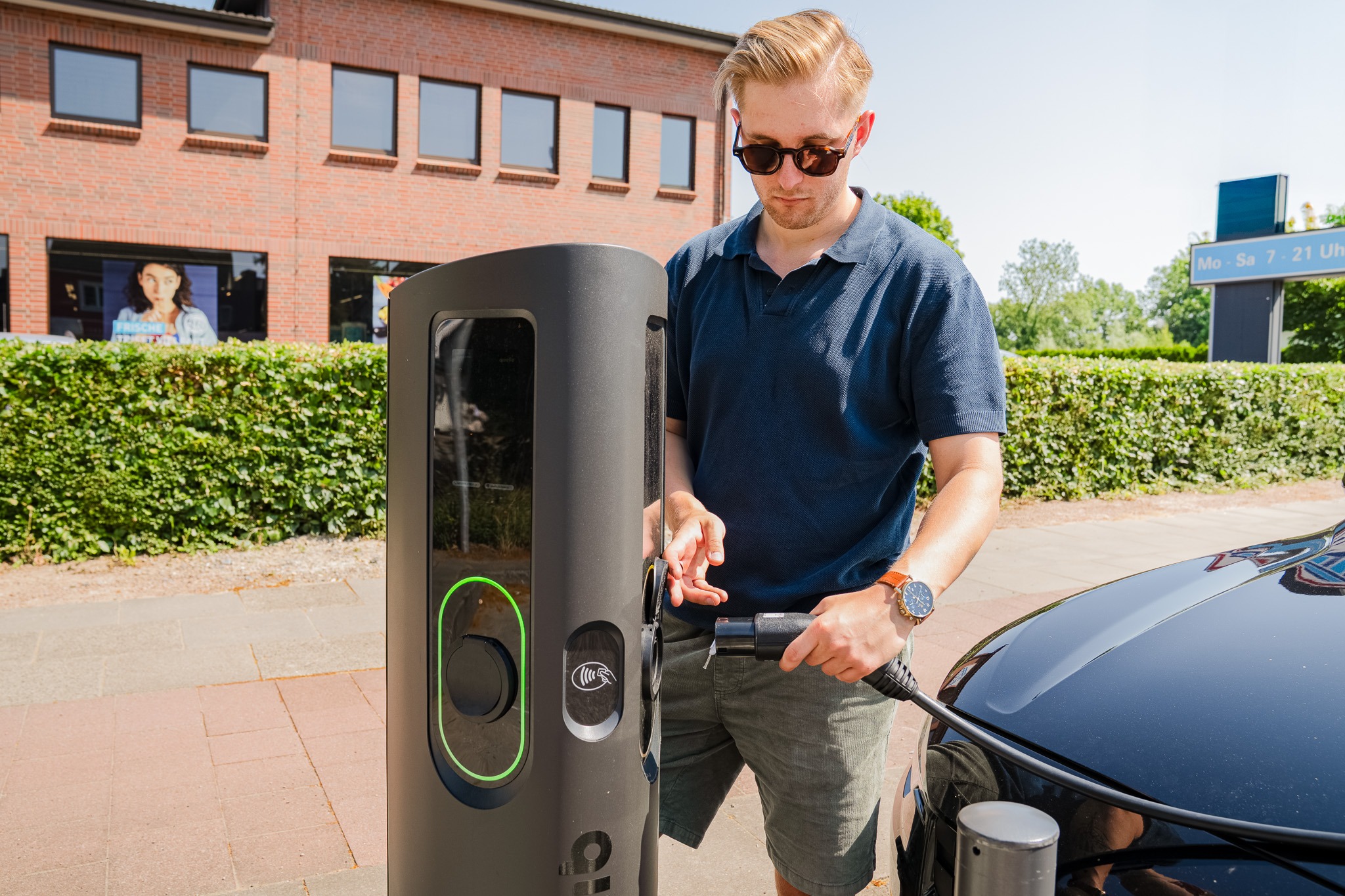 A man in a navy blue polo shirt plugging an EV charging cable into a public EV charging point. The cable also appears to be connected to his car which is partly visible in the background