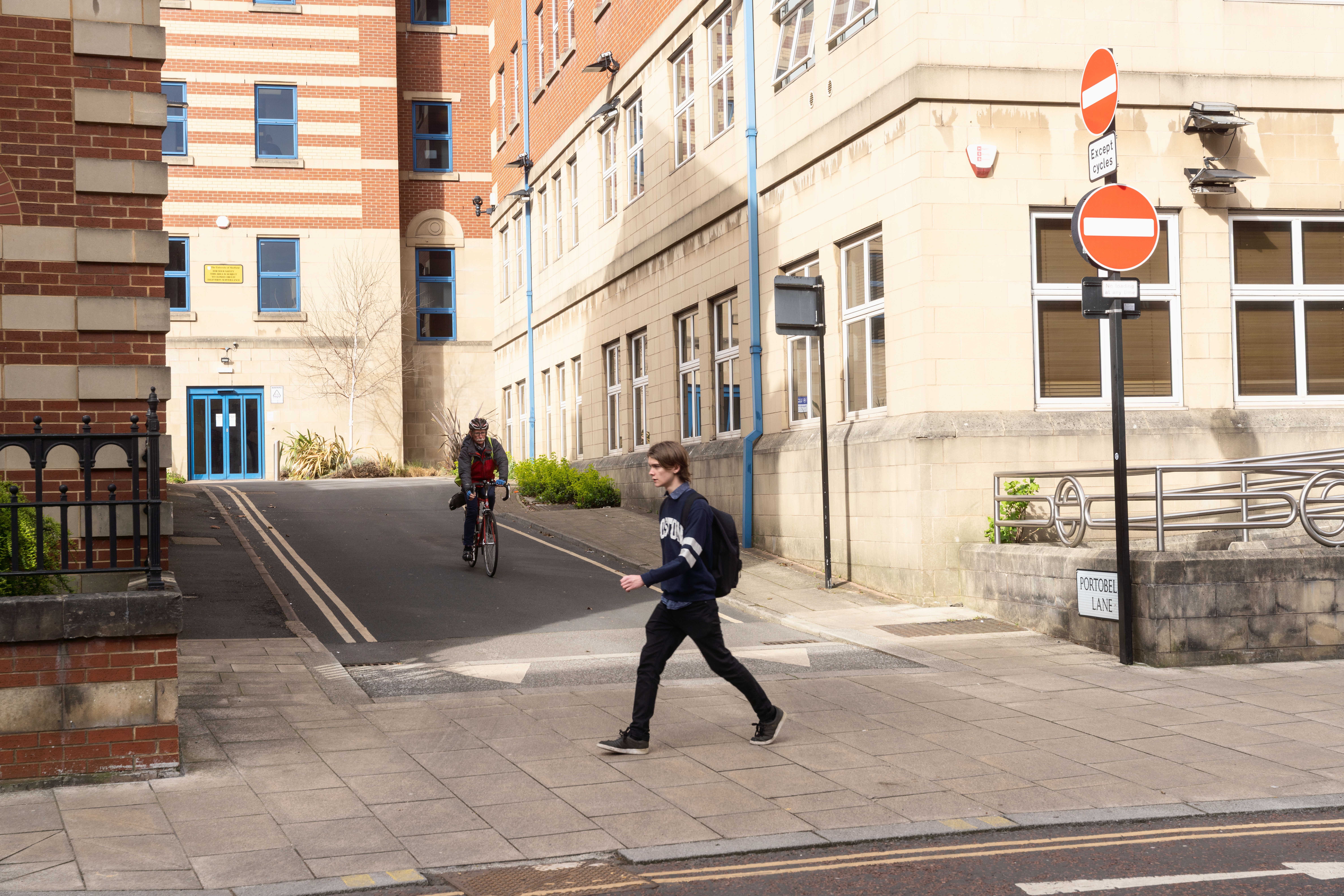 Image shows someone walking across a level pavement that crosses the junction of Mappin Street and Portobello Lane in Sheffield. A cyclist is approaching the junction from Portobello Lane. There are buildings in the background and a red and white No Entry sign to the right
