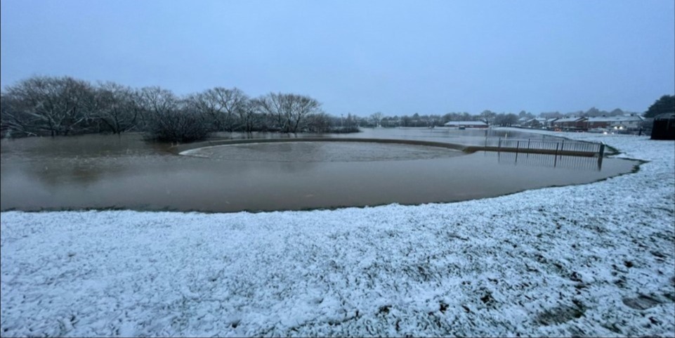 The same image as the previous one, albeit from a slightly different angle. However in this image there is snow on the ground the sky is a overcast and a gloomy white colour and the entire park is completely submerged in water a few metres deep. Water is flowing over the top of the circular spillway and flowing through a culvert under Dakyn road into the channel downstream.