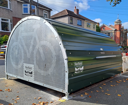 A green cucle hangar stands on a road with houses and cars in the background