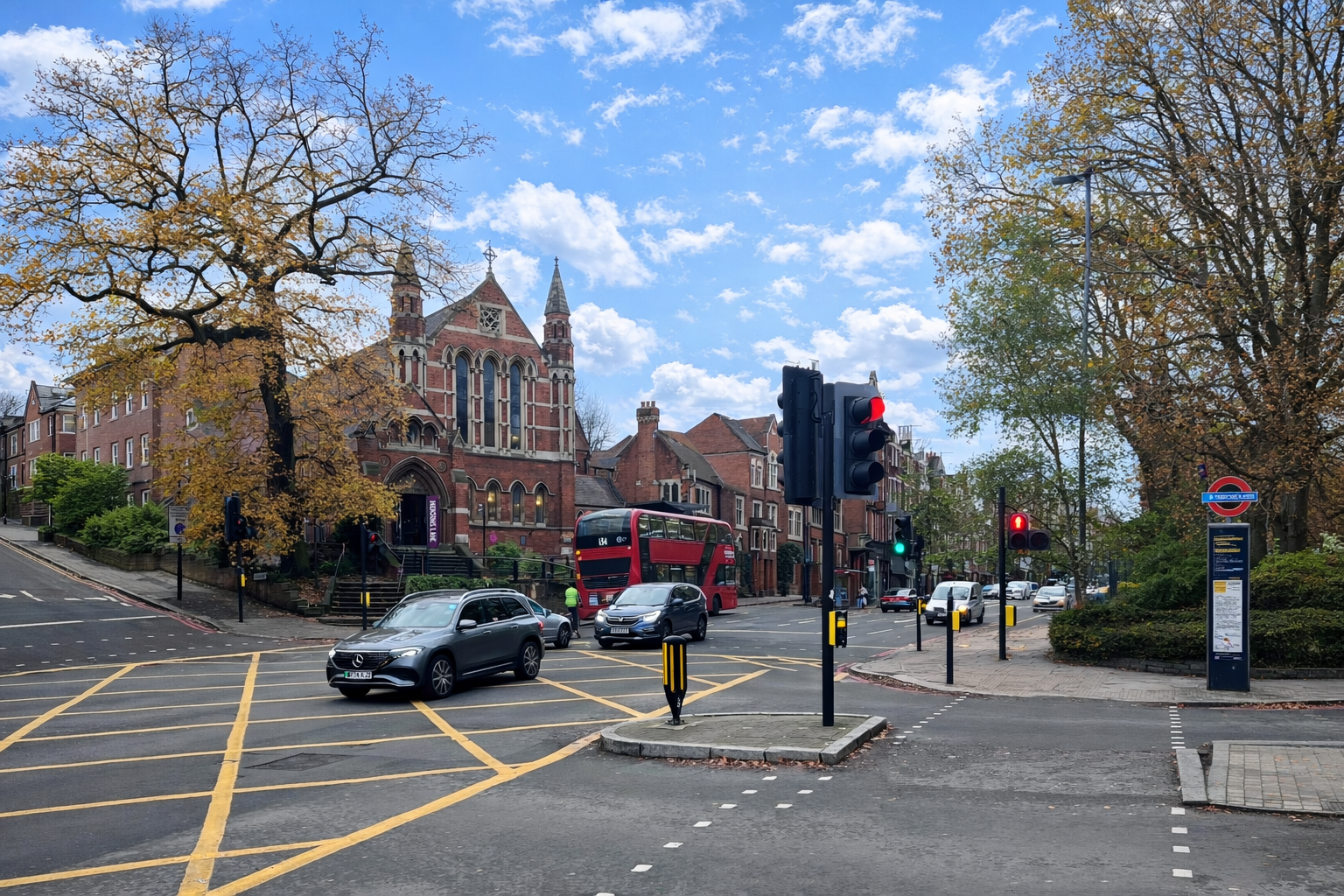 Shepherds Hill junction with yellow box markings, traffic lights, cars, and a red double‑decker bus beside a brick church and station sign under a blue sky