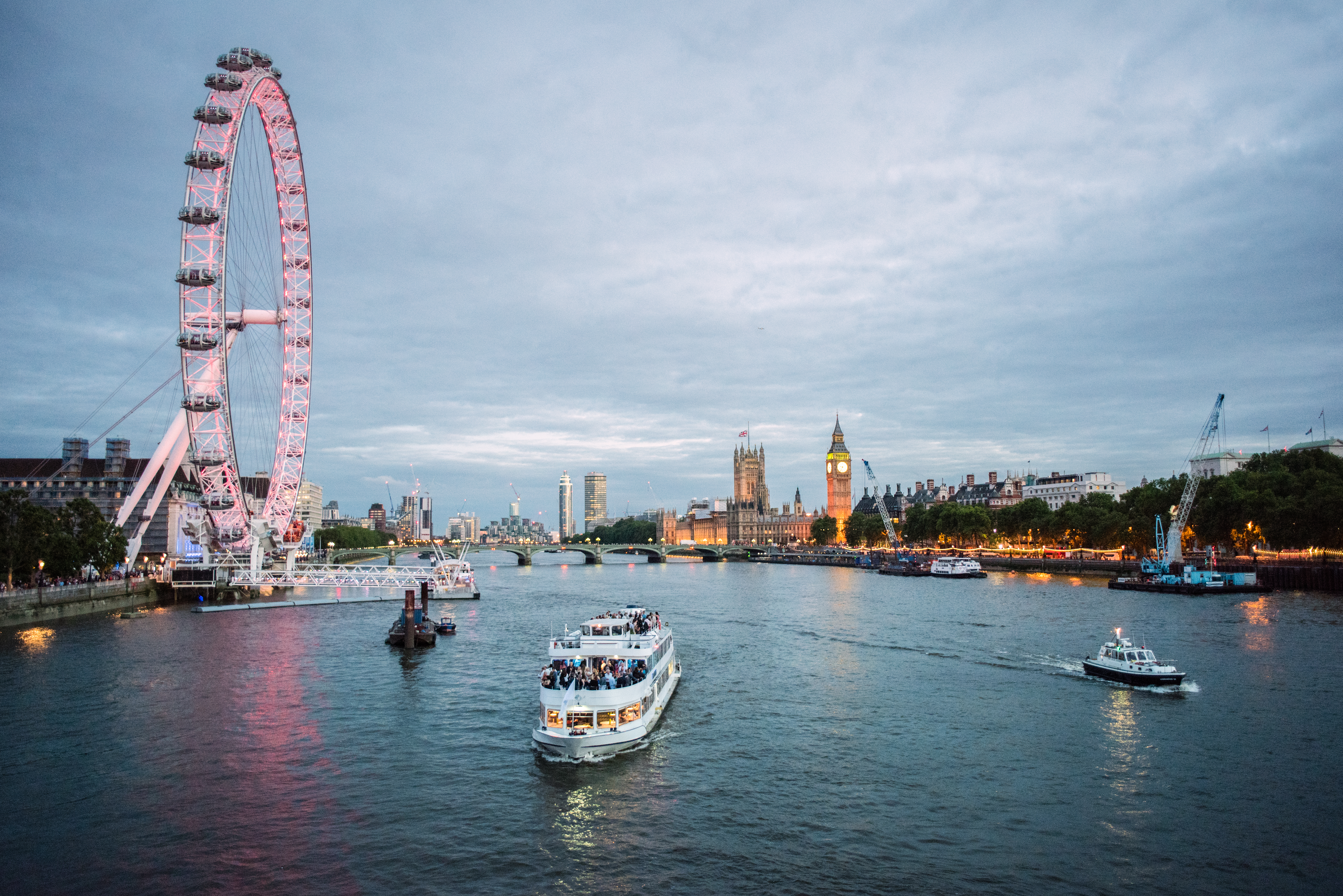 Evening view of the River Thames with the London Eye, Big Ben, and a boat cruising past illuminated city landmarks