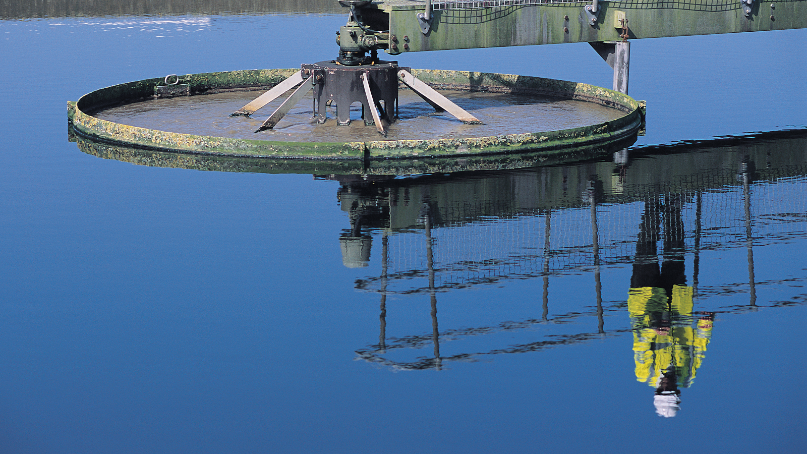 A circular water treatment tank with a rotating mechanism, reflecting a worker in a yellow safety suit against a calm blue water surface.